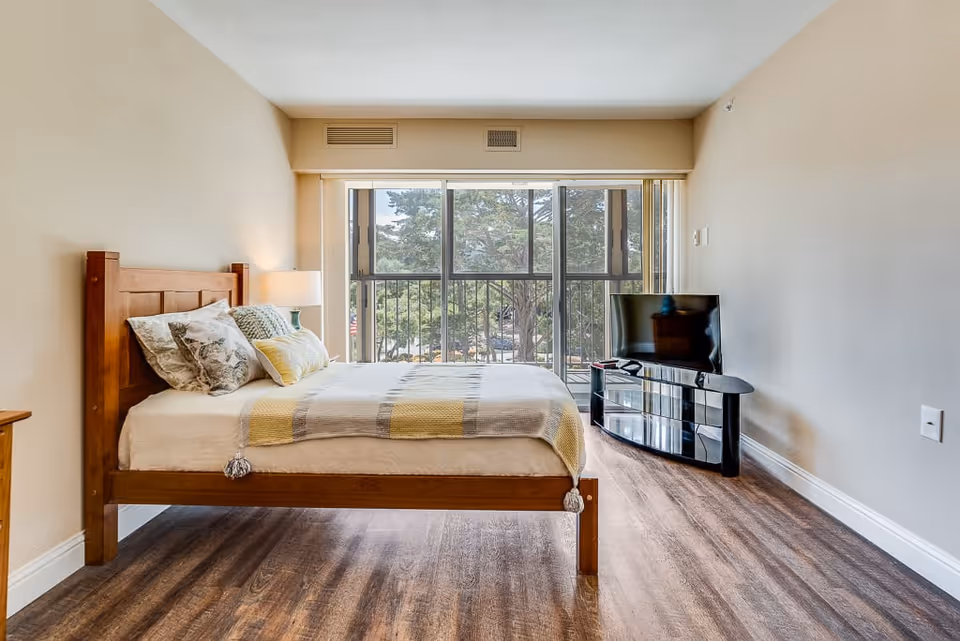 A bright bedroom with a wooden bed frame, neatly made bed with patterned pillows and a yellow and gray blanket. There is a bedside table with a lamp on the left side. A flat-screen TV sits on a black glass TV stand to the right. Large windows with a view of trees and a balcony railing allow natural light to fill the room. The floor is wood laminate and the walls are painted beige.
