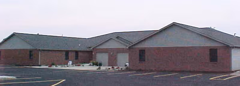 Exterior view of a single-story brick building with a gray roof, multiple garage doors, and a parking lot with marked spaces in front.