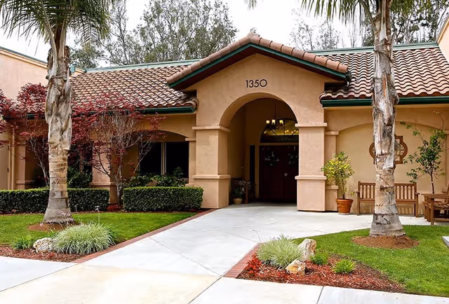 Senior living facility entrance with arched doorway, stucco walls, and manicured landscaping.