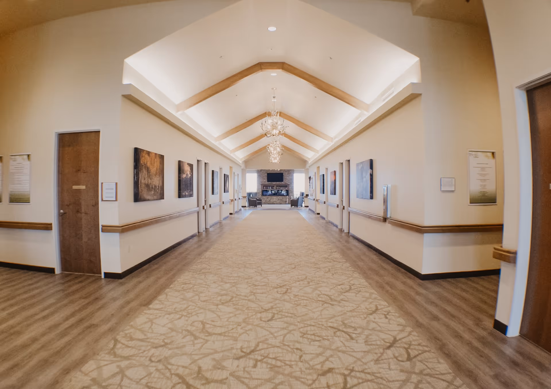 A wide, well-lit hallway with a high vaulted ceiling featuring wooden beams and modern chandeliers. The hallway has a patterned carpet runner in the center with wood flooring on the sides. Both walls are lined with closed wooden doors and framed artwork. At the far end, there is a seating area with chairs and a fireplace beneath a mounted television.