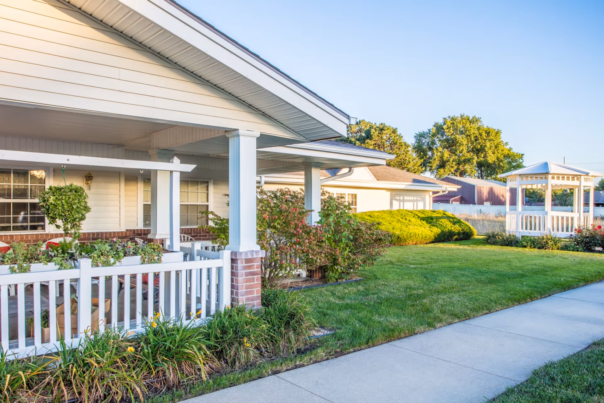Exterior view of a senior living facility with a covered porch, white railing, hanging plants, bushes, a well-maintained lawn, and a white gazebo in the background under a clear blue sky.
