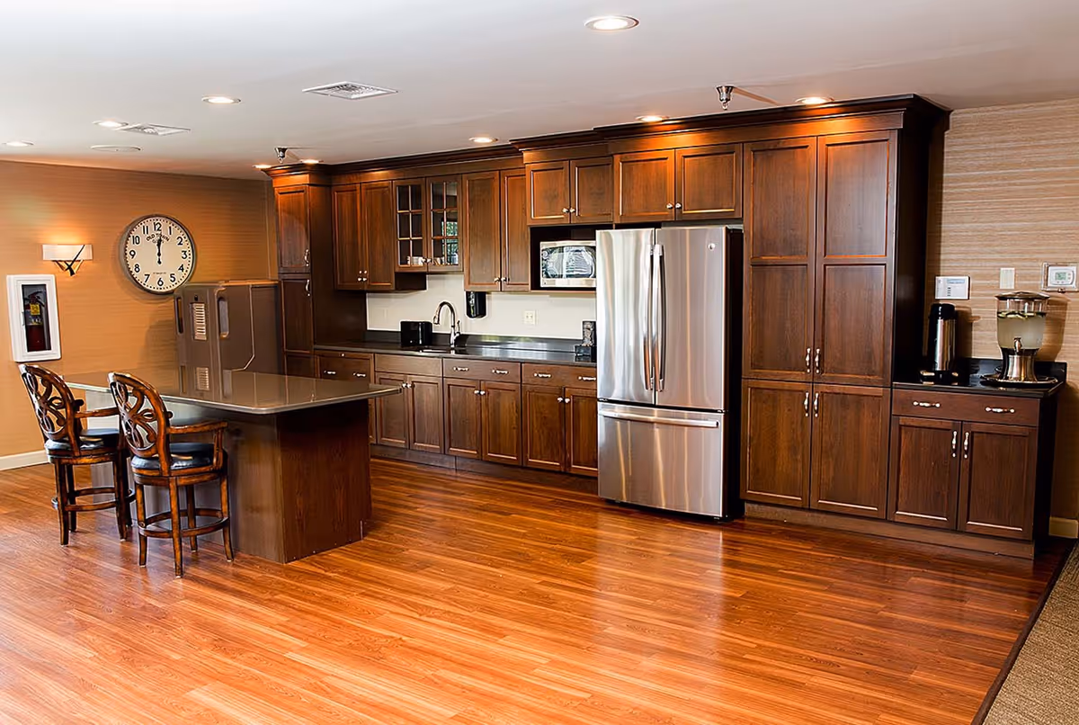 Communal kitchen with dark wood cabinets, stainless steel refrigerator, a center island with two chairs, and hardwood floors.