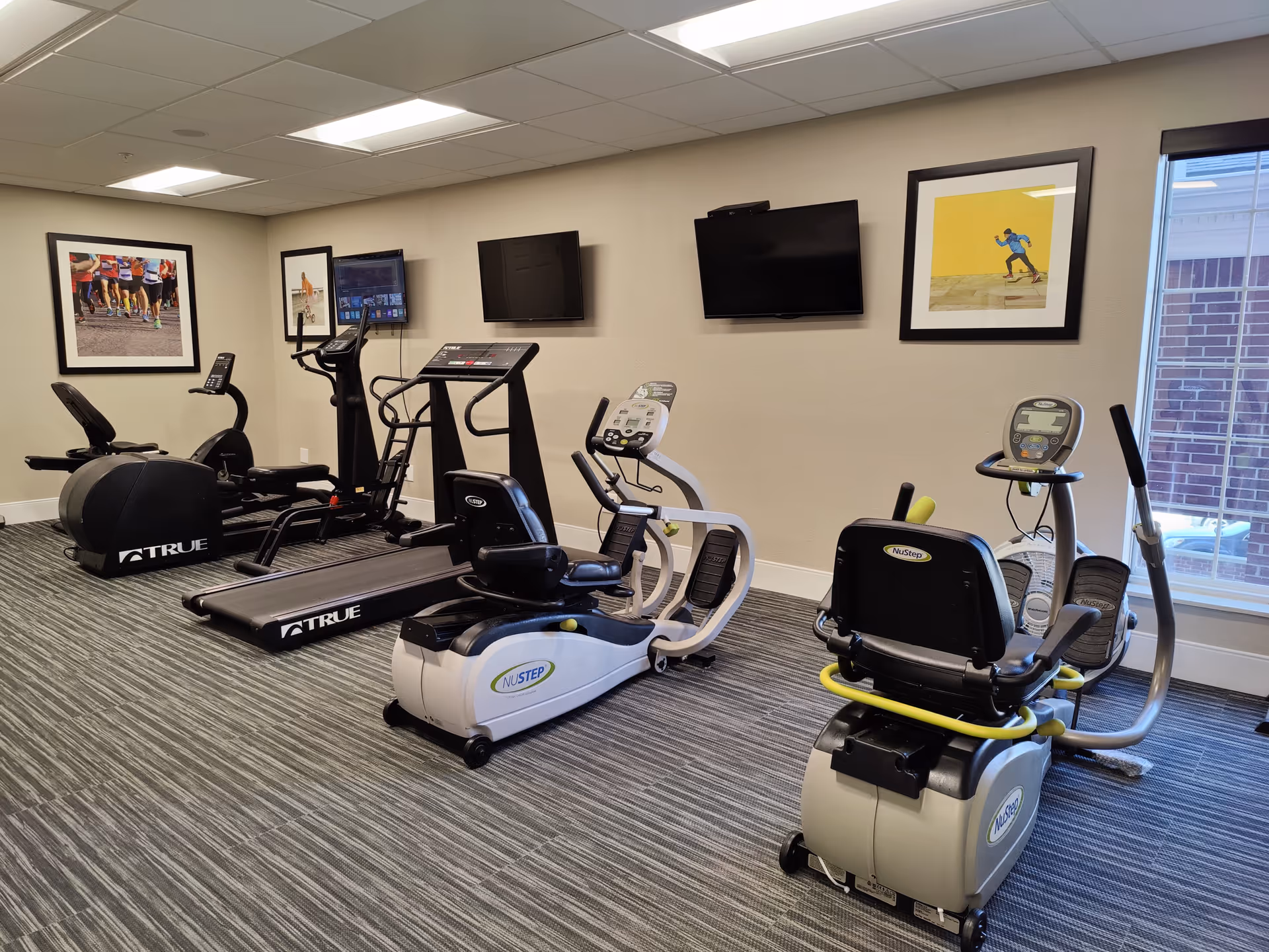 An exercise room with treadmills, recumbent bikes, ellipticals, wall-mounted TVs and framed artwork.