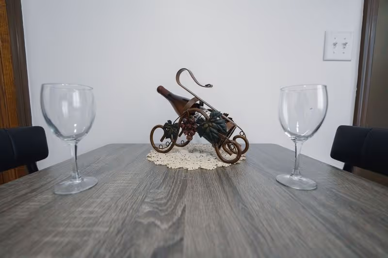 A wooden dining table with two empty wine glasses placed on either side. In the center of the table, there is a decorative wine bottle holder with a bottle, resting on a white lace doily. The background shows a plain white wall with a light switch and part of a wooden doorframe.