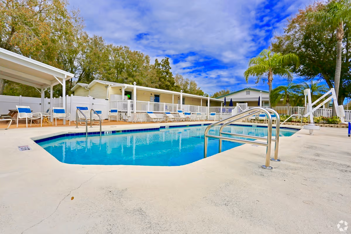 Outdoor swimming pool area with several lounge chairs around it, a white fence, trees, and a partly cloudy blue sky. There are buildings in the background and a pool lift for accessibility.