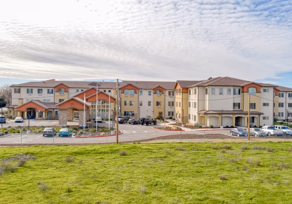 Exterior view of a large, multi-story senior living facility building with beige and orange accents, a parking lot with several cars, and a green grassy area in the foreground under a partly cloudy sky.