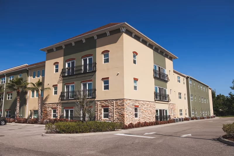 Exterior view of a three-story senior living facility building with beige and green walls, stone accents on the lower part, small balconies, and palm trees in front. The building is surrounded by a parking lot under a clear blue sky.