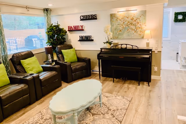 Light-filled common room with three leather recliners with green pillows, a piano against the wall, a pale blue coffee table and decorative wall art.