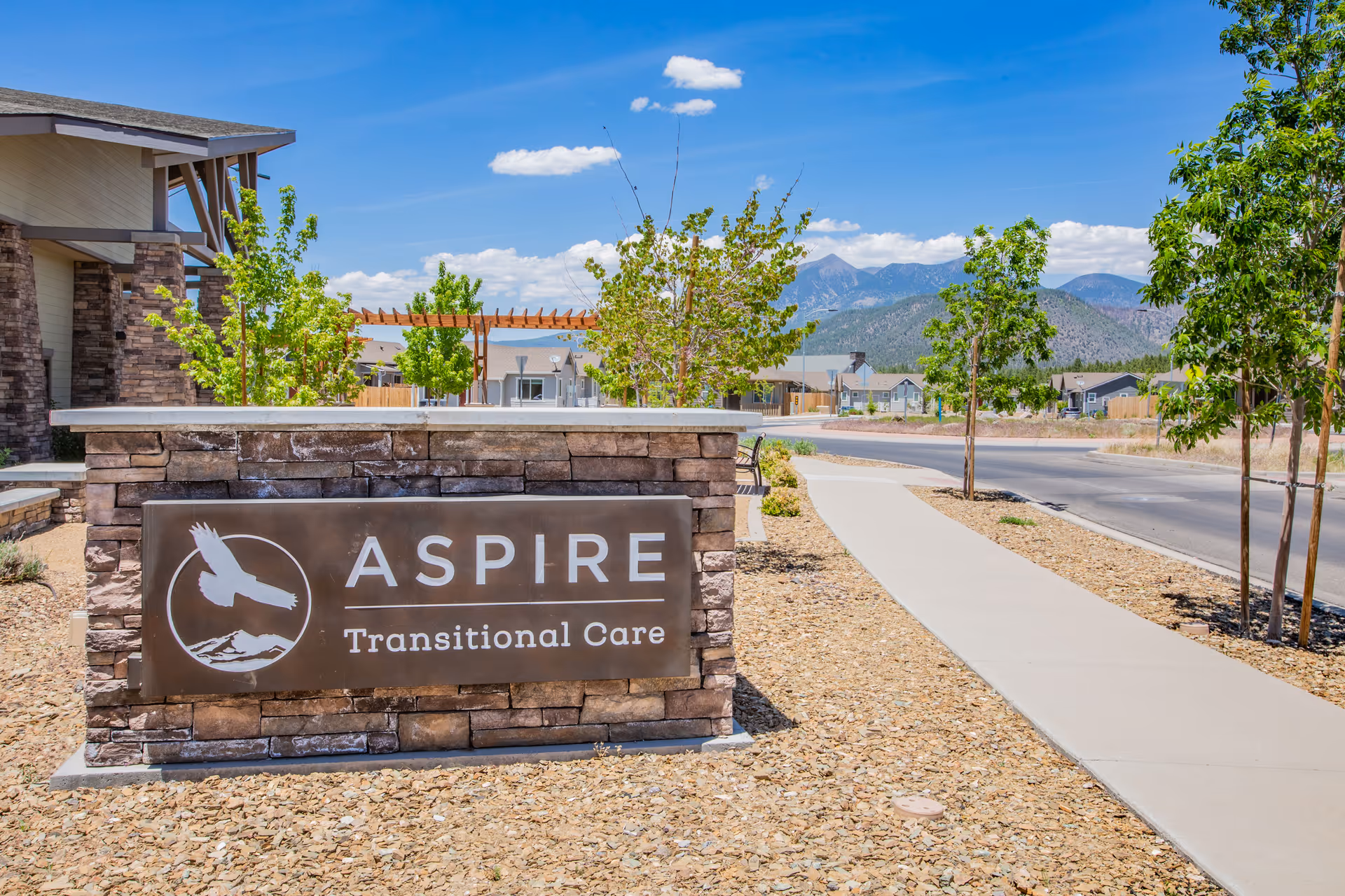 Outdoor view of Aspire Transitional Care facility sign made of stone with a metal plaque featuring a bird and mountain logo. The sign is placed on a landscaped area with small trees and a sidewalk leading towards the building under a blue sky with scattered clouds and mountains in the background.