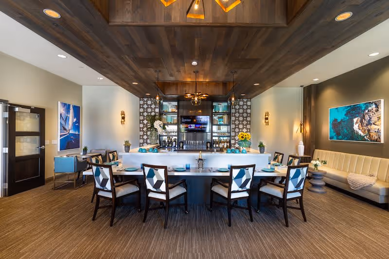 A modern dining area with a large rectangular table surrounded by chairs with geometric patterned cushions. The room features a bar area with shelves holding bottles and glasses, a ceiling fan, and decorative wall panels. There are framed pictures on the walls and a beige cushioned bench along one side. The floor is carpeted and the ceiling has wooden paneling with recessed lighting.