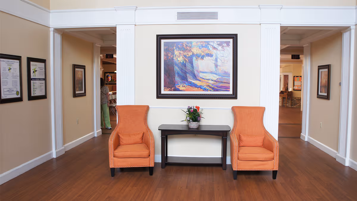 A hallway area in a senior living facility with two orange armchairs placed on either side of a small dark wooden table holding a flower arrangement. Above the table hangs a framed colorful painting depicting a forest scene with sunlight streaming through trees. The walls are painted beige with white trim and molding. There are framed pictures and notices on the walls along the hallway, and a person is partially visible in the background walking through a doorway.