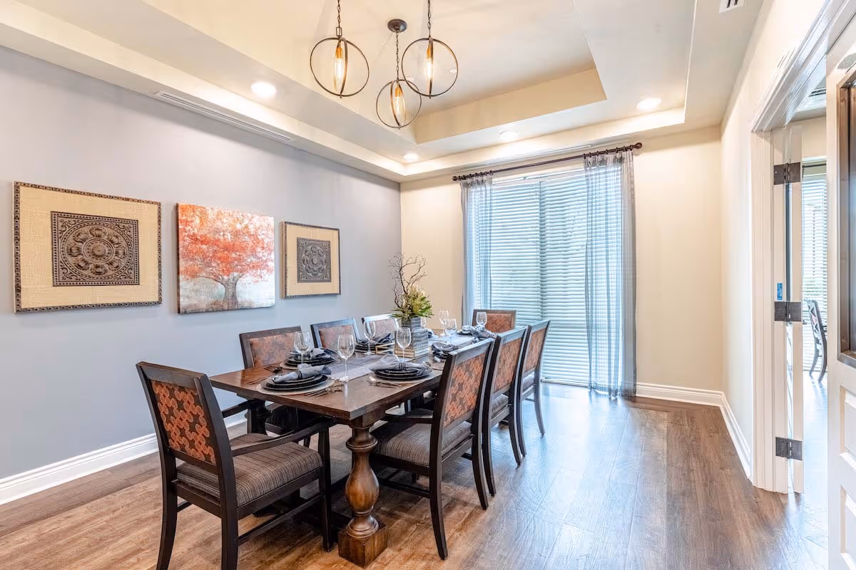 A well-lit dining room with a wooden dining table set for eight people. The table has plates, napkins, and wine glasses arranged neatly. There are three framed artworks on the light gray wall, including a painting of a tree in the center. The room features wooden flooring, a large window with blinds and sheer curtains, and three modern pendant lights hanging from the ceiling.