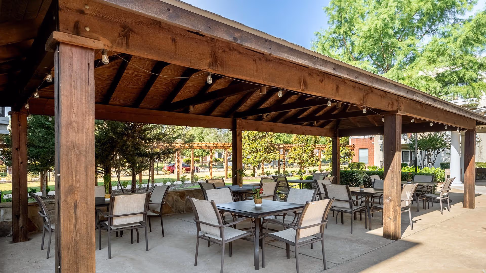 Outdoor covered patio area with multiple tables and chairs arranged for seating. The wooden structure has a slanted roof with string lights hanging underneath. Surrounding the patio are trees, shrubs, and a view of a garden area with a pergola in the background.