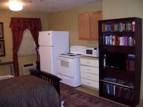 Studio-style patient room with bed, refrigerator, stove, microwave, and bookshelf.