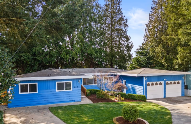 Exterior view of a single-story blue house with white trim, surrounded by tall trees and a well-maintained lawn with shrubs and a small tree in front. The house features a two-car garage with white doors and a driveway leading up to it.