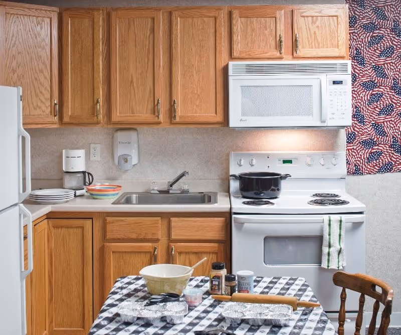 A kitchen with wooden cabinets, a white refrigerator, a white stove with a black pot on it, and a white microwave above the stove. There is a sink with a faucet in the countertop, a coffee maker, plates, and bowls on the counter. A table with a black and white checkered tablecloth holds baking supplies including a mixing bowl, rolling pin, spices, and foil pans. An American flag-themed curtain is visible on the right side.
