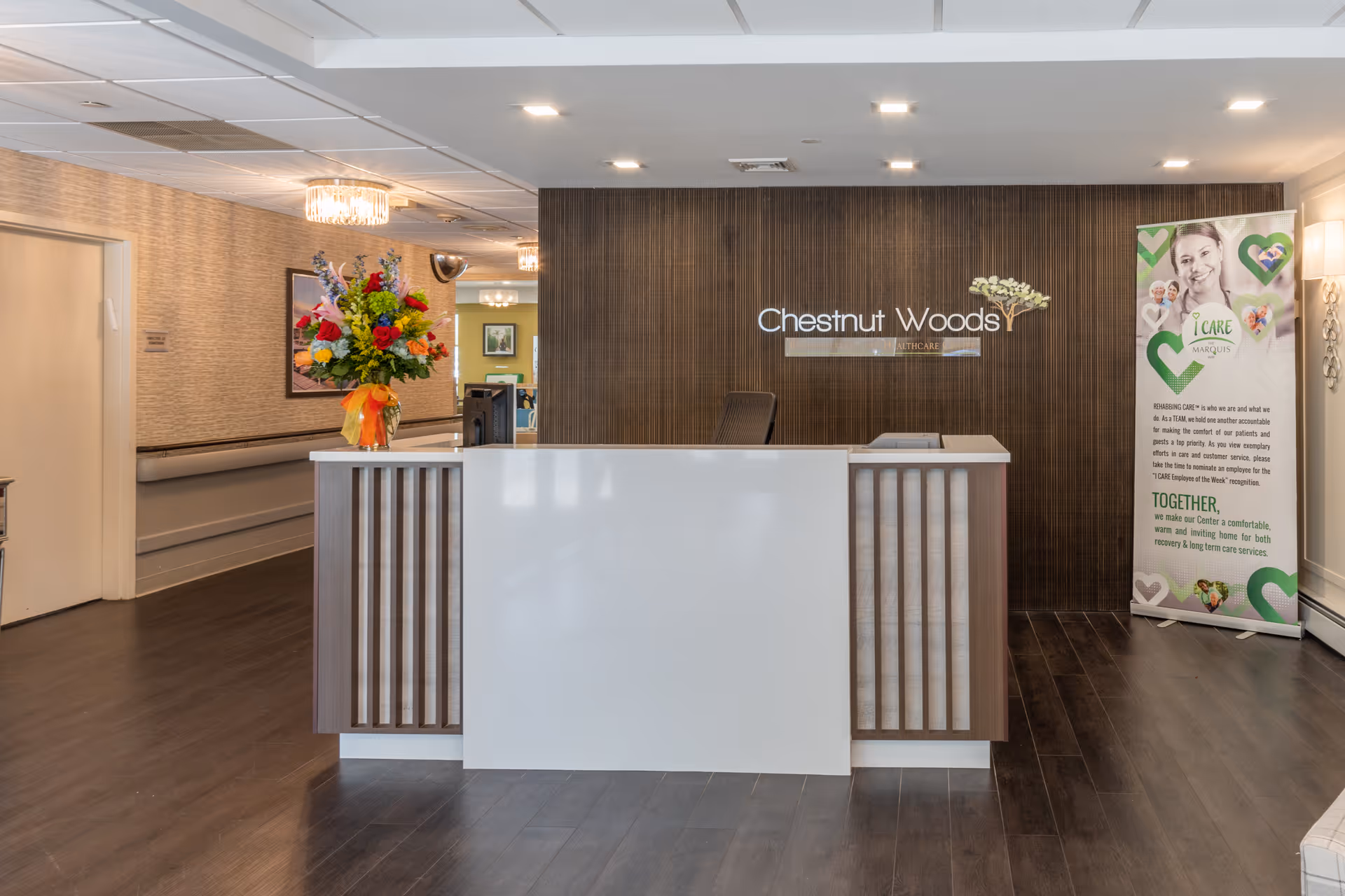 Reception area of Chestnut Woods Rehabilitation & Healthcare Center featuring a modern white and wood front desk with a colorful flower arrangement on the left side. Behind the desk is a dark wood accent wall with the facility's name displayed. To the right, there is a standing banner with information and images related to care services. The floor is dark wood, and the ceiling has recessed lighting.