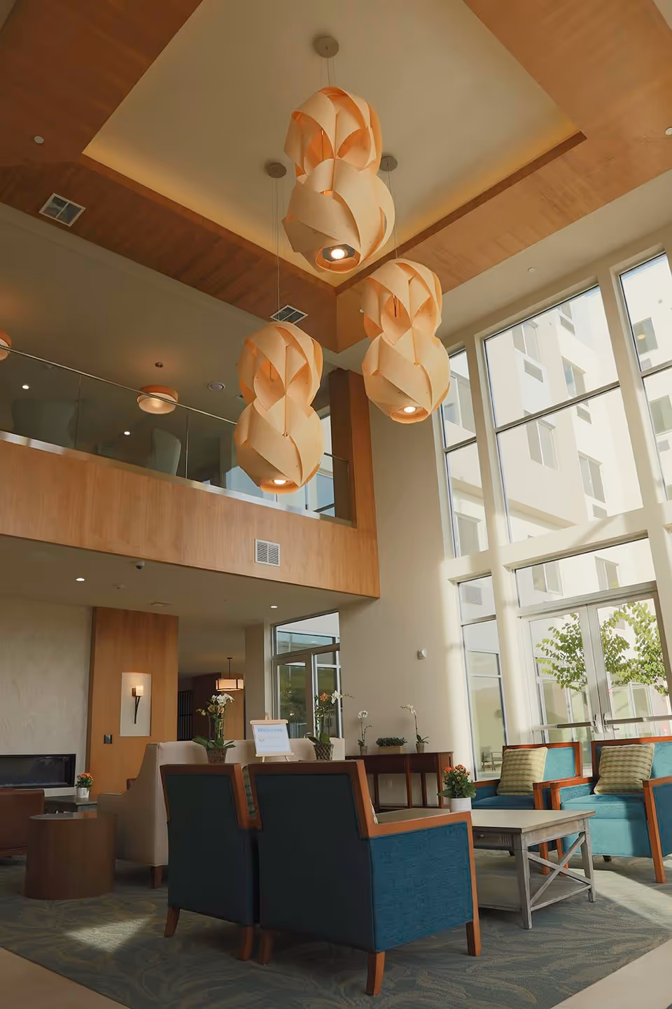 A spacious and well-lit senior living facility common area with high ceilings, large windows, and modern hanging light fixtures. The room features comfortable seating with blue and beige chairs arranged around a coffee table, decorative plants, and a fireplace in the background. A balcony with glass railing overlooks the area.