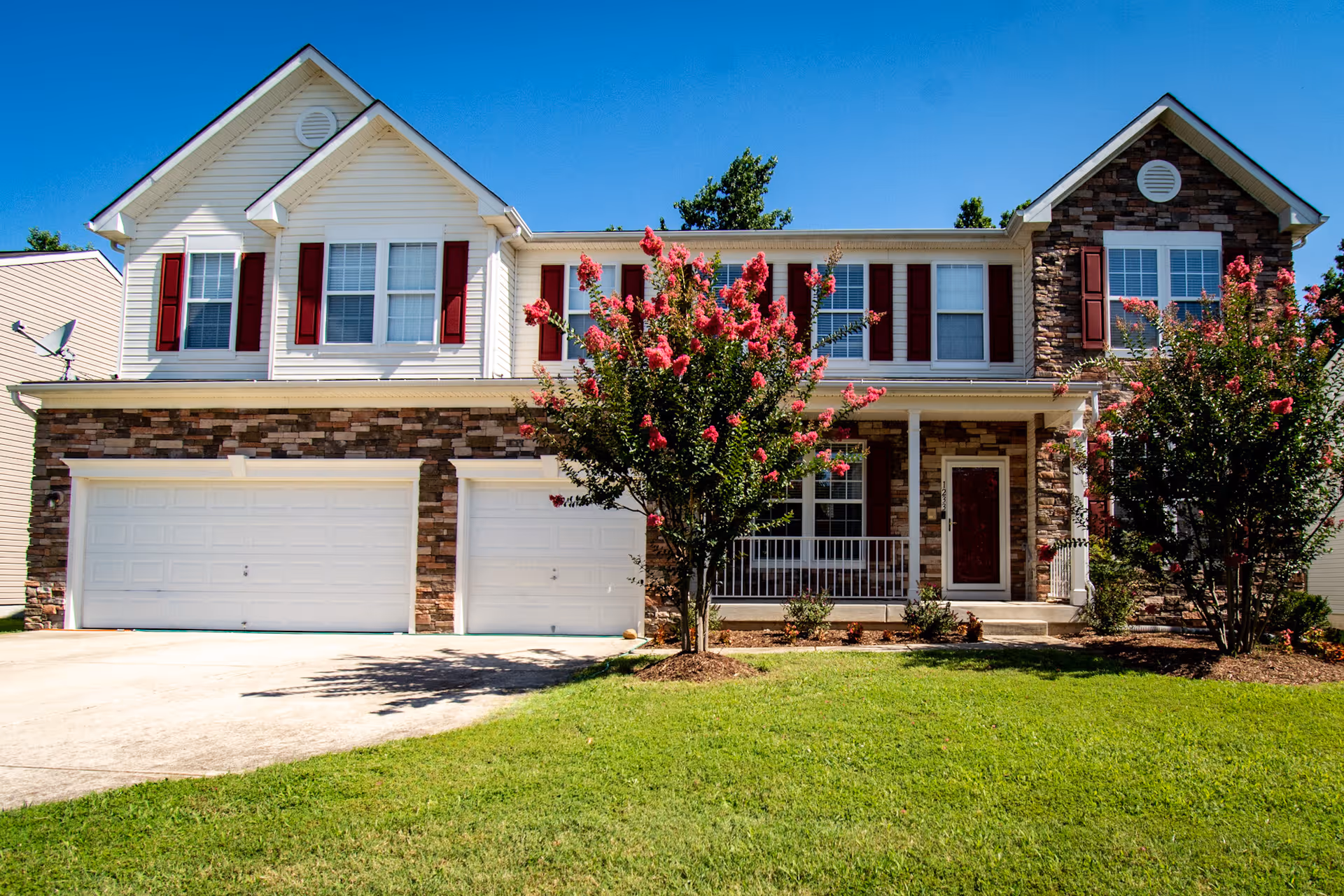 Front exterior view of a two-story residential building with white siding and stone accents, red shutters, a three-car garage, a porch with white railing, and flowering trees in the front yard under a clear blue sky.