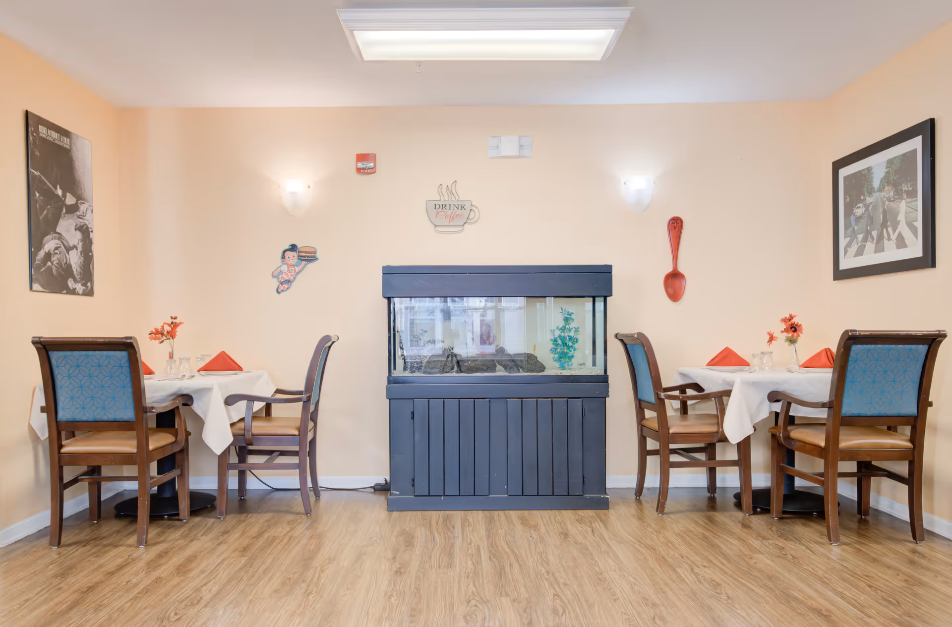 A cozy dining area with two tables, each set with white tablecloths, red napkins, and small flower vases. Four wooden chairs with blue patterned cushions surround each table. A large aquarium with a black stand is centered against a peach-colored wall, decorated with wall art including a large red spoon, a coffee cup sign, and framed pictures. The floor is light wood, and the room is softly lit by wall sconces and a ceiling light.