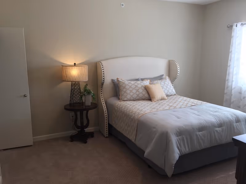 A neatly made bedroom with an upholstered headboard, patterned bedding, a bedside table with a lamp and plant, and a curtained window.