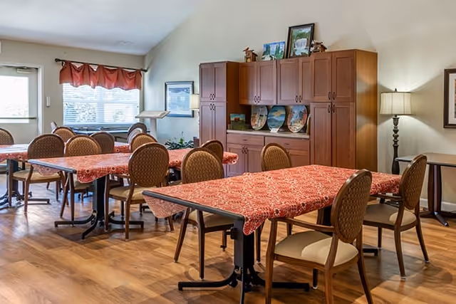 A dining room with multiple tables covered in red patterned tablecloths and surrounded by brown cushioned chairs. The room has wooden flooring, a large wooden cabinet with decorative plates and framed pictures on top, a floor lamp, and windows with red valance curtains letting in natural light.