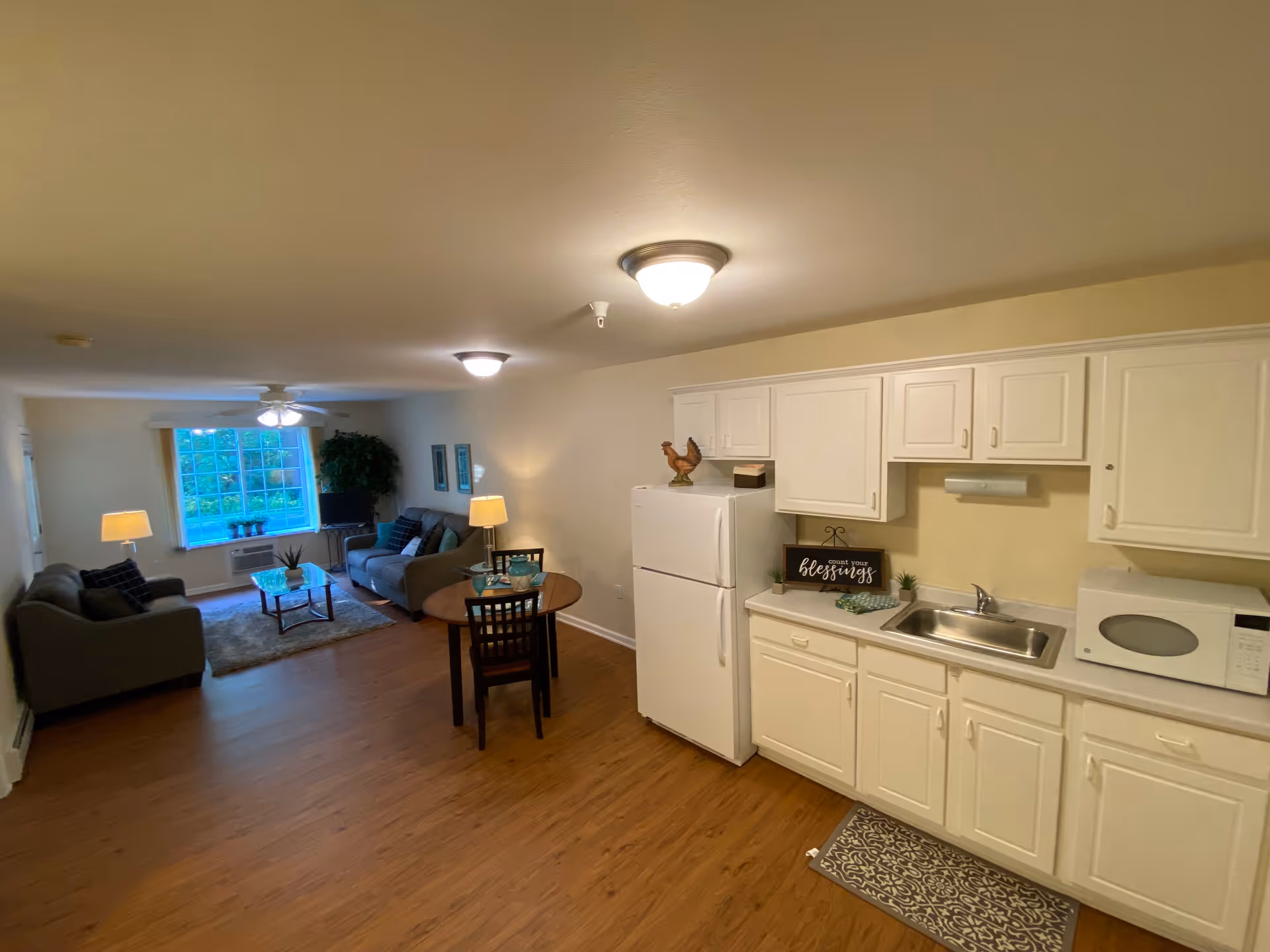 Interior view of a senior living facility apartment showing a small kitchen area with white cabinets, a refrigerator, microwave, and sink on the right. A round dining table with two chairs is in the center. In the background, there is a living room area with two sofas, a coffee table, lamps, a large window, and a ceiling fan.