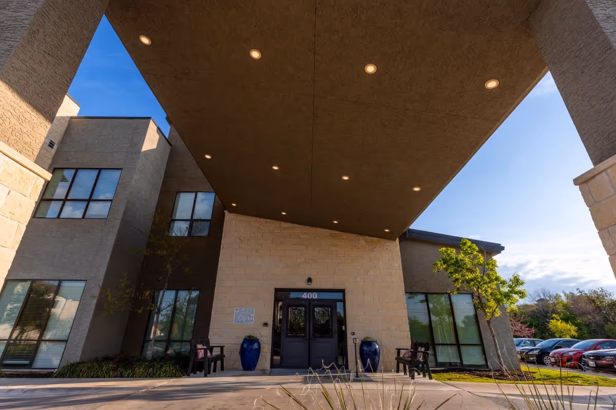 Entrance of a modern building with a covered driveway, large windows, two blue decorative pots, benches on either side of the door, and a parking lot with cars visible to the right. The building has a beige and brown exterior with a clear blue sky above.