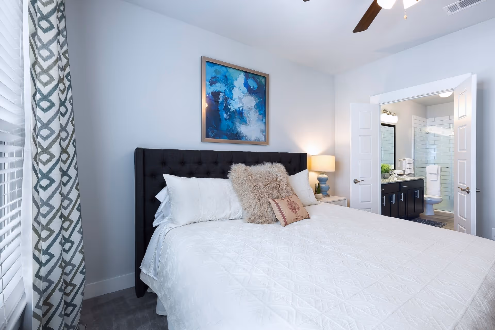 A bedroom with a large bed featuring a dark tufted headboard, white bedding, and decorative pillows. A nightstand with a lamp is beside the bed. A framed abstract blue and white painting hangs on the wall above the bed. To the right, double doors open to reveal a bathroom with a vanity, mirror, toilet, and a glass-enclosed shower. A window with patterned curtains is on the left side of the room.