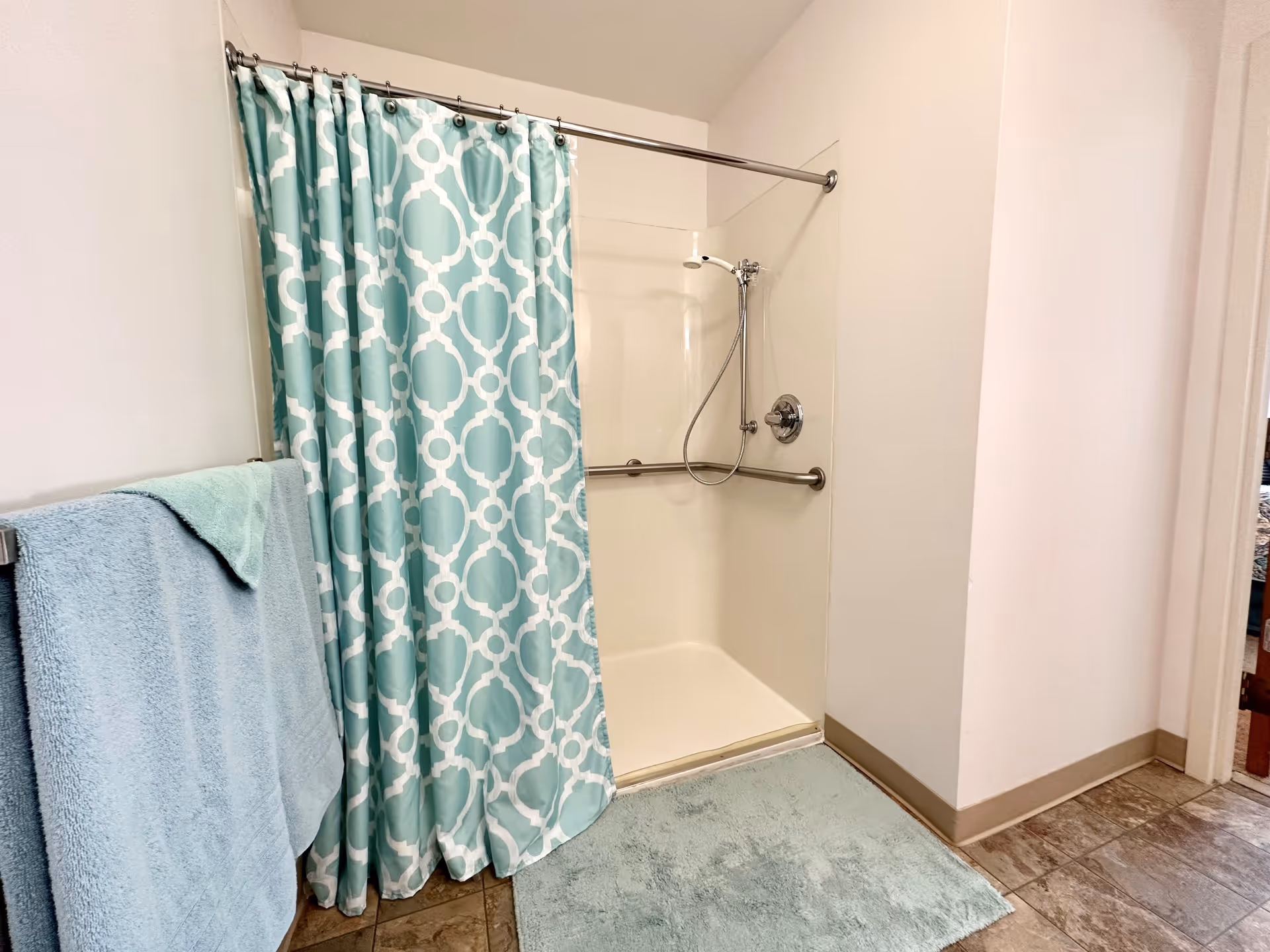 Shower stall with a teal patterned curtain, grab bars, handheld showerhead, and matching towels and bath mat.
