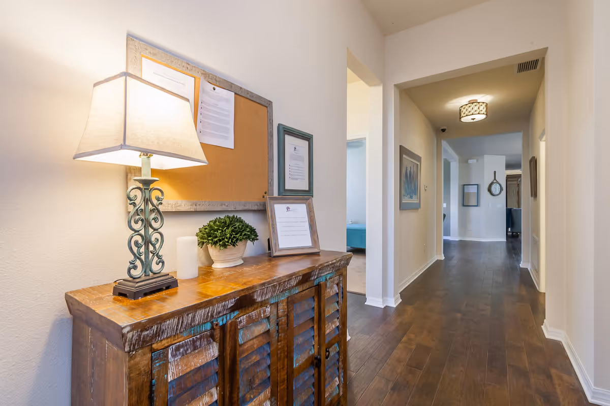 A well-lit hallway with wooden flooring and light-colored walls. On the left side, there is a rustic wooden cabinet with a decorative lamp, a small potted plant, and framed documents on top. A cork bulletin board with papers is mounted on the wall above the cabinet. The hallway leads to other rooms, with a glimpse of a blue bed in one room and a wall clock visible further down the hall.