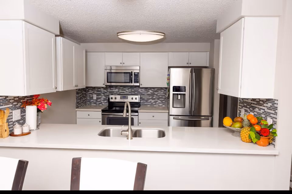 A modern kitchen with white cabinets, a stainless steel refrigerator, microwave, and stove. The countertop is white with a double sink and a silver faucet. There are decorative items including a bowl of fruit and flowers on the counter. The backsplash features a mosaic tile design in shades of gray.