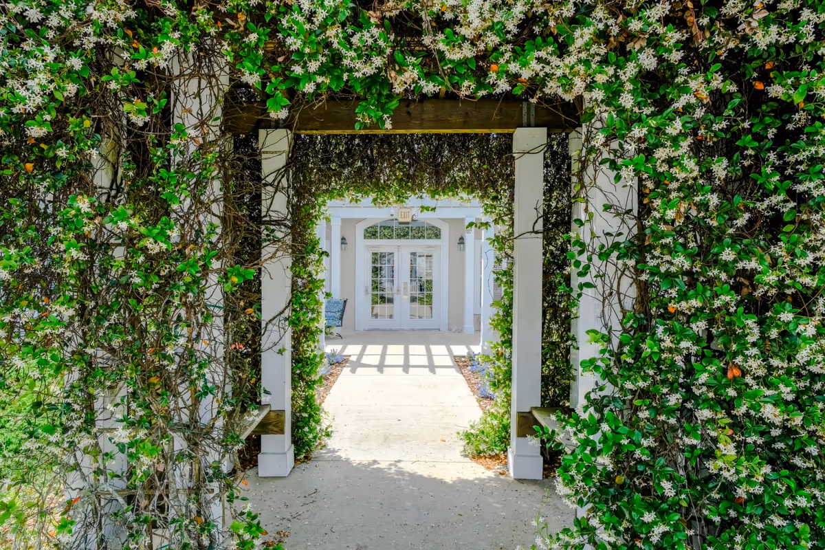 A walkway framed by a white pergola covered in green vines and white flowers leading to a building entrance with double glass doors and an exit sign above.