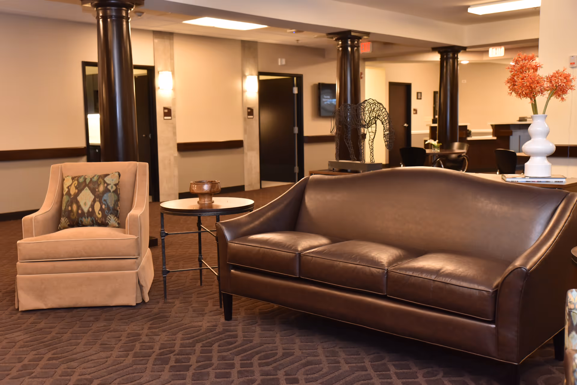 A comfortable seating area in a senior living facility featuring a brown leather sofa, a beige armchair with a patterned cushion, a small round side table, and decorative items including a wireframe horse sculpture and a white vase with orange flowers. The background shows a hallway with dark wooden columns and doors.