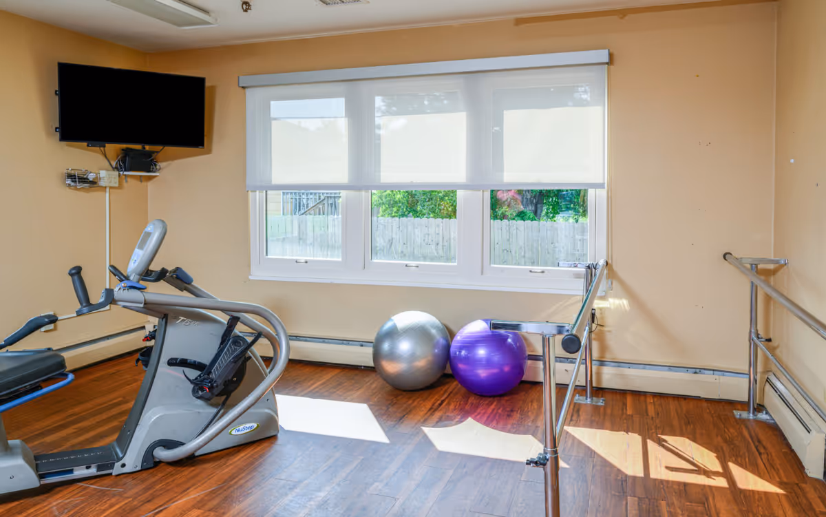 Indoor fitness room with a treadmill, exercise balls, a ballet barre, a wall-mounted TV, and a window with blinds.