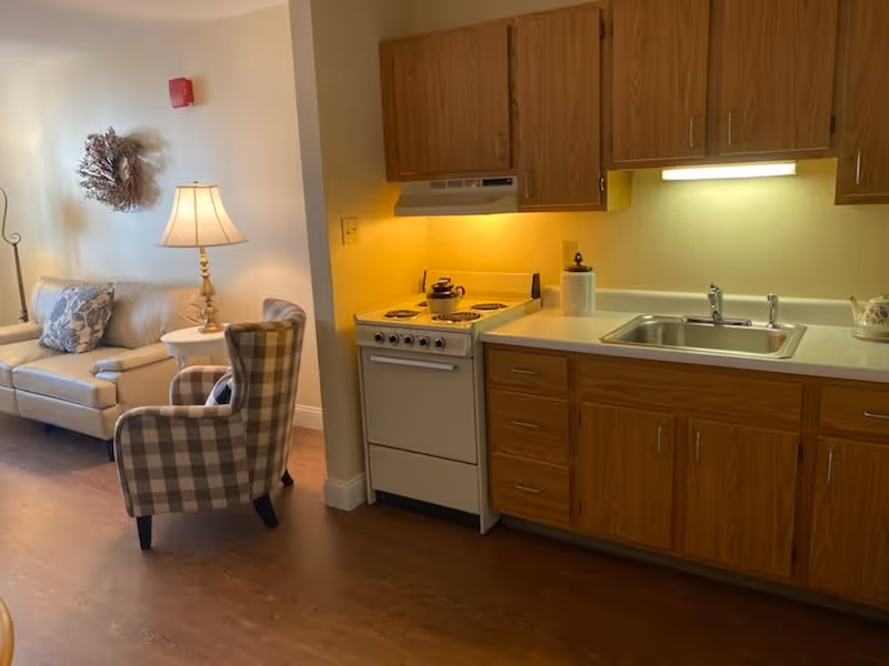 A cozy interior space featuring a small kitchen area with wooden cabinets, a white stove, and a sink. Adjacent to the kitchen is a living area with a beige sofa, a plaid armchair, a side table with a lamp, and a decorative wreath on the wall.