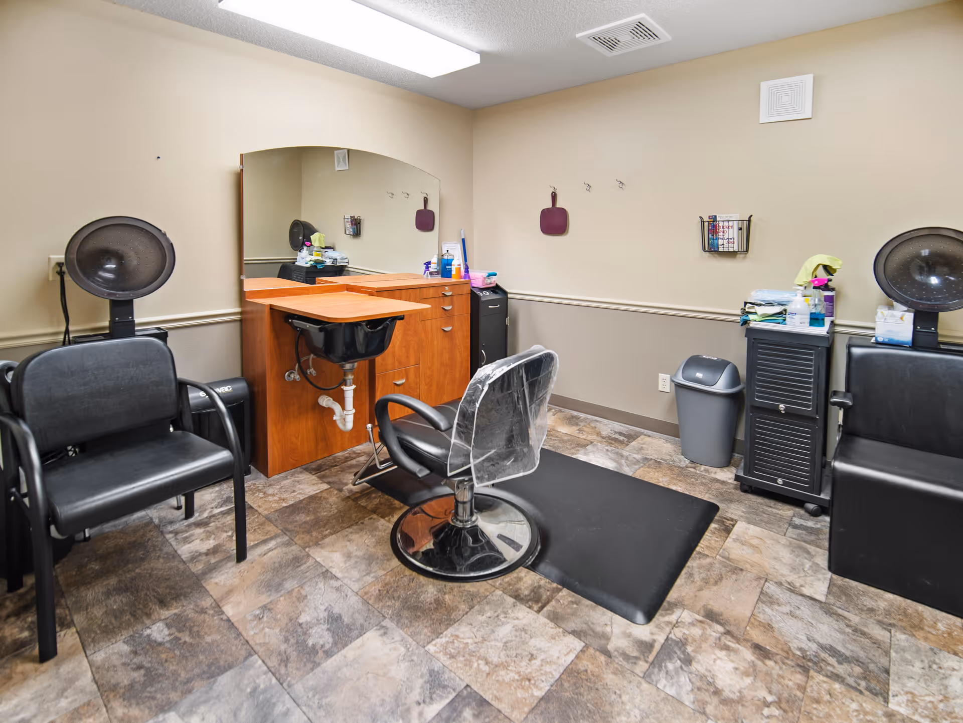 Interior view of a small hair salon area with two black salon chairs, two hair dryers, a wooden cabinet with a sink, a large mirror on the wall, and various hair care products and supplies. The floor has a stone tile pattern, and the walls are painted beige with a chair rail molding.