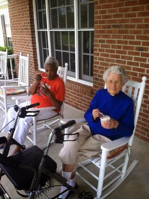 Two elderly women sitting on white rocking chairs on a covered porch with a brick wall behind them. One woman is wearing a red shirt and white pants, eating from a bowl, and the other woman is wearing a blue sweater and beige pants, also holding a bowl and spoon. A walker is positioned in front of the woman in the red shirt.