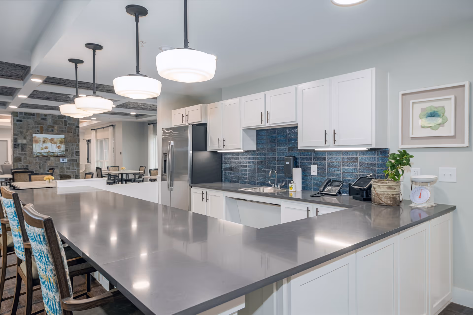 Modern kitchen area in a senior living facility with a long gray countertop, white cabinets, stainless steel refrigerator, blue tiled backsplash, and pendant lights hanging from the ceiling. There are chairs with patterned cushions along the counter and a small potted plant and kitchen scale on the counter.