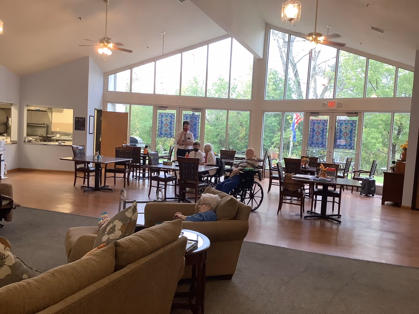 A spacious dining and common area in a senior living facility with large windows letting in natural light. Several elderly residents are seated at tables and in wheelchairs, with one person standing and interacting with them. The room has high ceilings with ceiling fans and a view of greenery outside. Comfortable seating with couches is visible in the foreground.