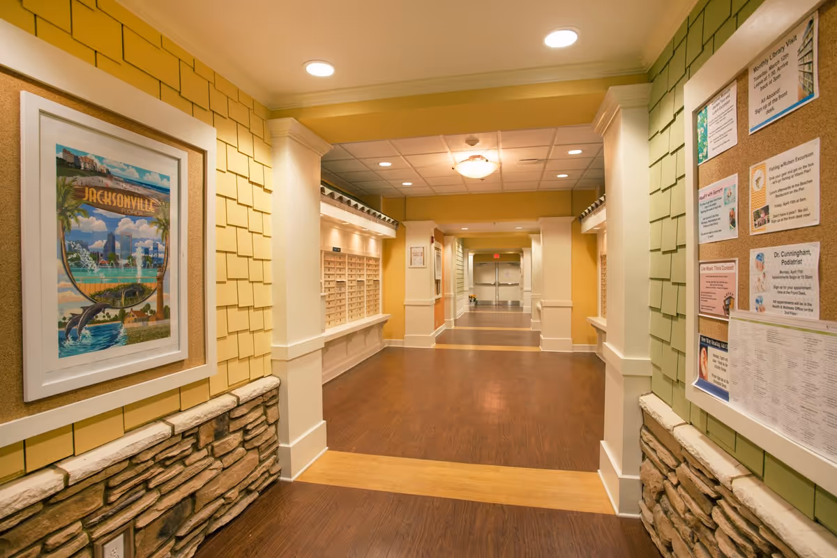 Interior hallway of a senior living facility with yellow and green walls, wood flooring, a framed Jacksonville artwork on the left wall, and a bulletin board with notices on the right wall. The hallway leads to double doors at the far end and has mailboxes along the left side.