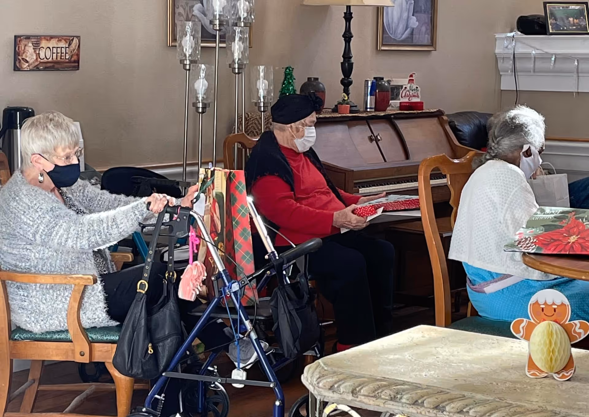 Three elderly individuals wearing face masks are seated in a cozy room. One woman with white hair and a gray sweater is sitting on a wooden chair with a walker in front of her. A man in a red shirt and black hat is seated near a piano, holding a wrapped gift. Another woman with gray hair and a white shawl is sitting at a table with holiday-themed decorations. The room has warm lighting, a lamp, framed artwork, and a sign on the wall that says 'COFFEE'.