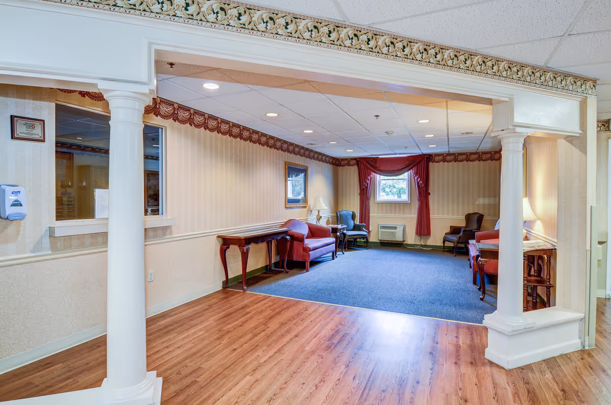 A cozy sitting area in a senior living facility with wooden flooring in the foreground and blue carpet in the seating area. The room features a red loveseat, two armchairs, side tables with lamps, a window with red curtains, and decorative molding along the ceiling. Two white columns frame the entrance to the seating area.