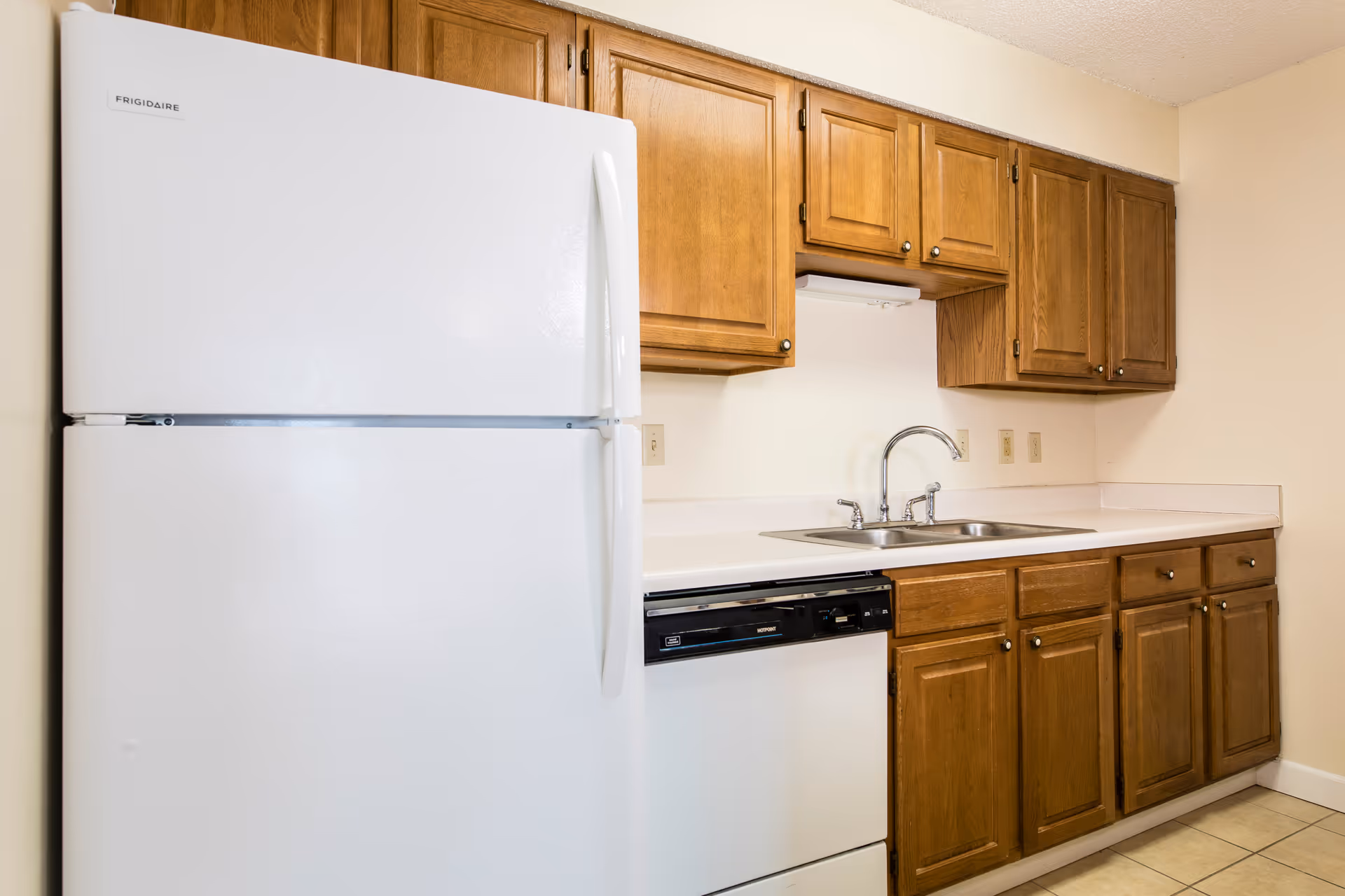 A kitchen area featuring a white Frigidaire refrigerator, wooden upper and lower cabinets, a double stainless steel sink with a chrome faucet, a white countertop, and a built-in dishwasher with a black control panel. The walls are light-colored and the floor is tiled.