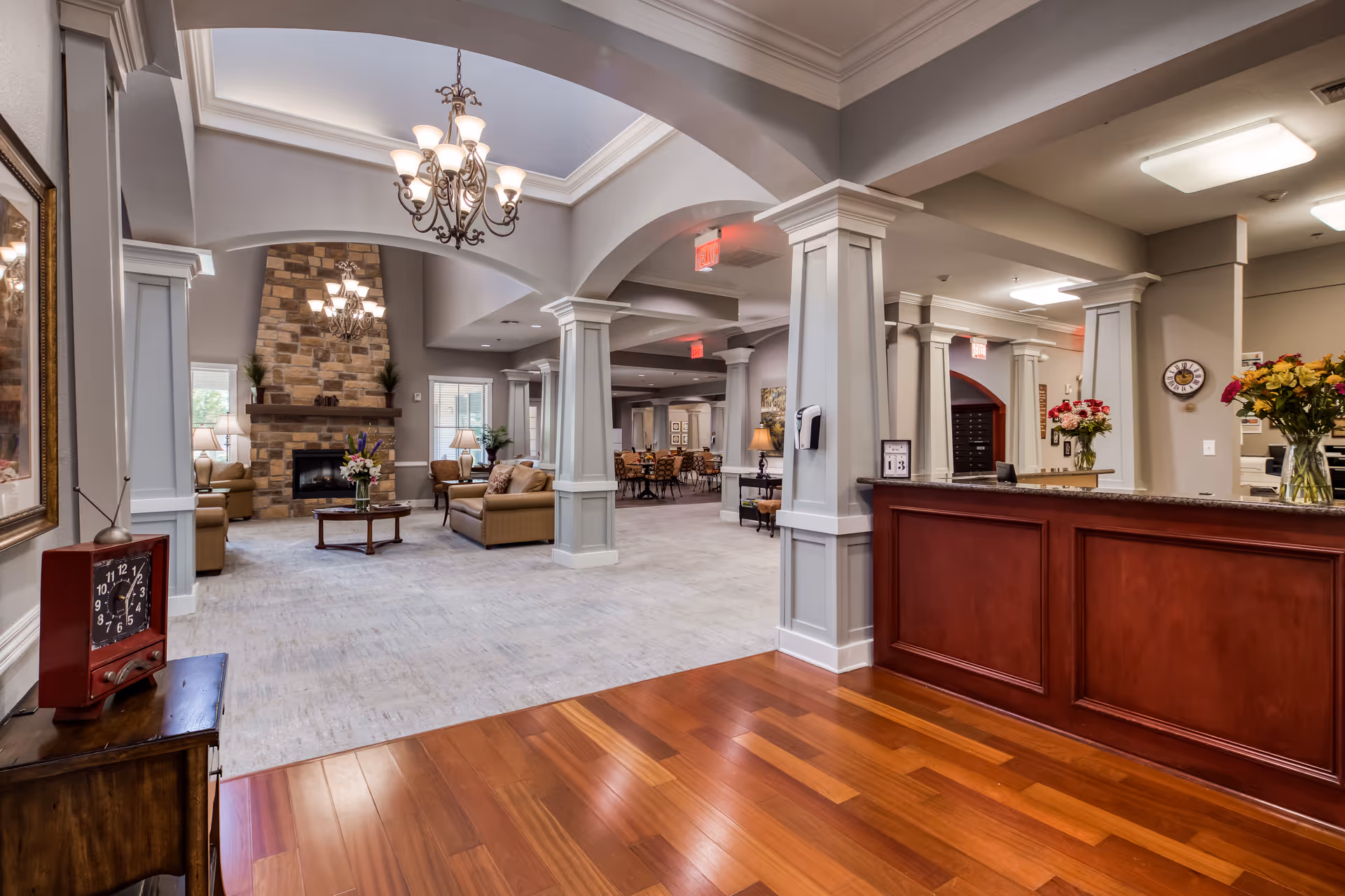 Spacious and elegant assisted living facility common area with a wooden reception desk on the right, a seating area with sofas and a coffee table in front of a large stone fireplace, decorative columns, chandeliers, and a dining area in the background.