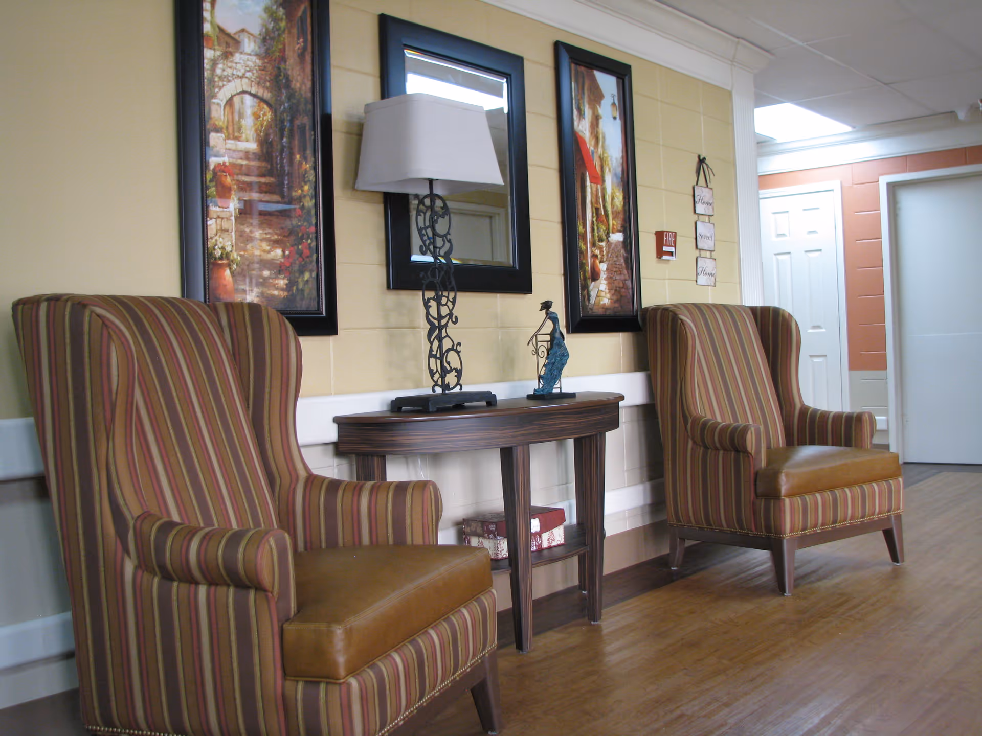 A cozy seating area with two striped armchairs facing each other, separated by a wooden console table with a decorative lamp and a small sculpture. The wall behind features two framed paintings and a mirror. The floor is wooden, and there are doors and a hallway visible in the background.