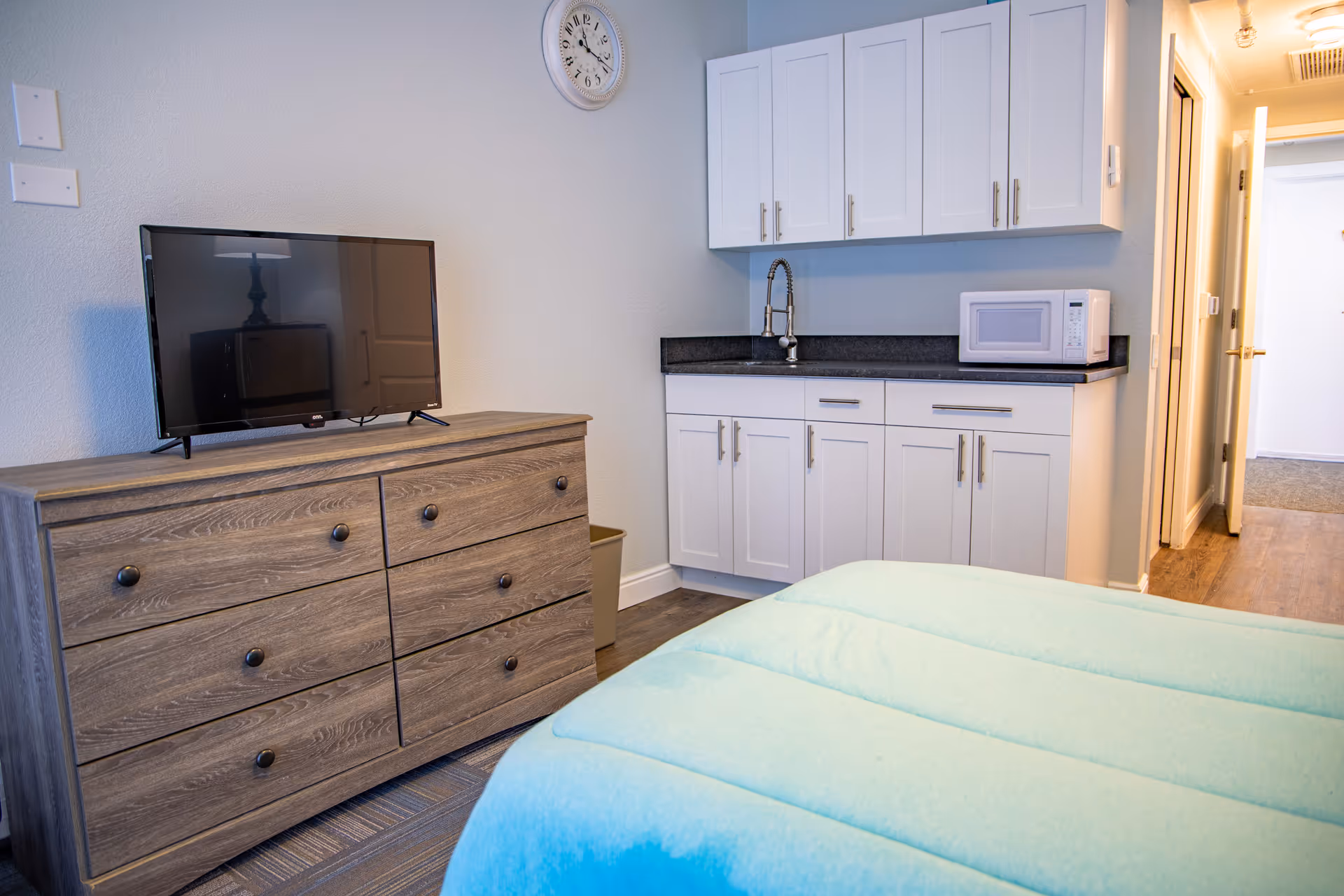 A senior living facility room featuring a wooden dresser with a flat-screen TV on top, a kitchenette area with white cabinets, a sink, and a microwave, and a bed with a light blue blanket. A clock is mounted on the wall above the dresser, and a hallway is visible in the background.