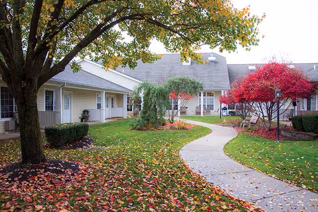 Curved concrete walkway through a landscaped courtyard with fall foliage and single‑story residential buildings.