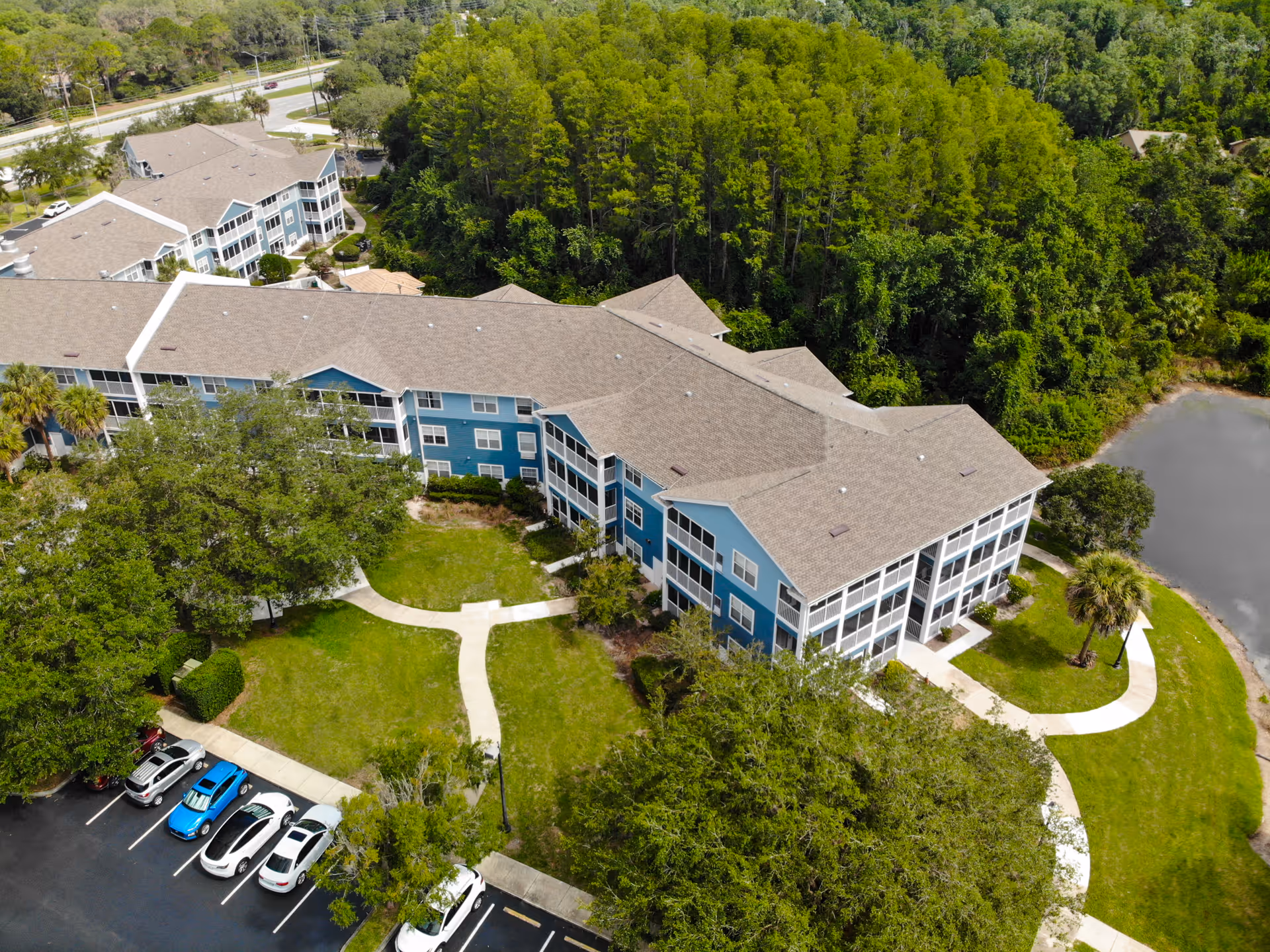 Aerial view of Addington Place of East Lake, showing a large multi-story residential building with blue siding and white trim surrounded by green trees and grass. There are paved walkways, a parking lot with several cars, and a small pond near the building.