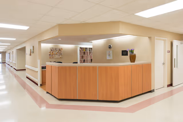 A clean and well-lit reception desk area in a healthcare or rehabilitation facility. The desk is made of light wood with a beige countertop, and there is a vase with purple flowers on it. Behind the desk are shelves with files and a wall clock. The hallway adjacent to the desk has handrails and extends into the distance with multiple doors along the corridor.